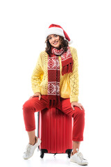 joyful young woman in santa hat and scarf with ornament sitting on travel bag isolated on white