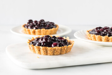 Straight on view of blueberry tarts on white plates and a white marble slab against a white background. 