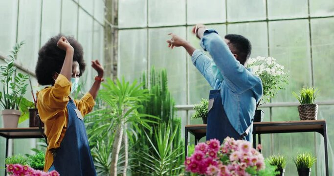 Portrait of happy African American male and female workers giving high five and dancing in flower shop at work. Cheerful woman and man florists in maskshaving fun. Employment concept