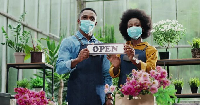 Portrait of joyful African American male and female in masks standing in flower shop and holding Open sign. Woman and man florists workers looking at camera in good mood. Family business concept