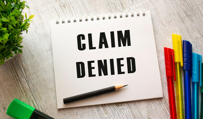 A notebook on a spring with the text CLAIM DENIED lies on a wooden table with colored pens.