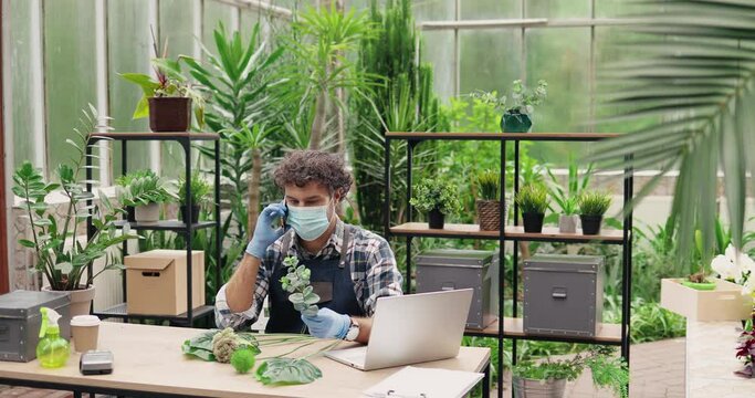 Young concentrated Caucasian male worker of flower shop sitting at table and calling on smartphone. Handsome man garden center owner in mask working and talking on cellphone. Florist concept