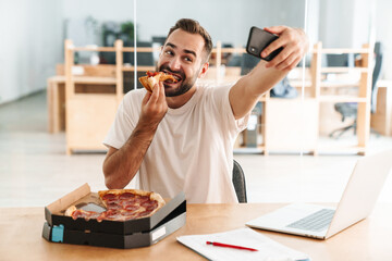 Caucasian joyful man eating pizza while taking selfie on cellphone