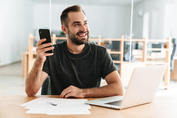 Happy man smiling and using cellphone while working with laptop