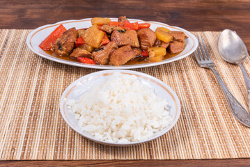 Adobe pork with pineapple and bell pepper in an oval plate and a bowl of rice on a bamboo napkin - philippine cuisine