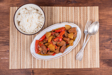 Adobe pork with pineapple and bell pepper in an oval plate and a bowl of rice on a bamboo napkin - philippine cuisine