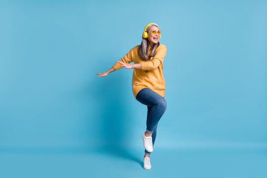 Can't be bothered. Full length photo of charming dancer aged lady listen music earphones hands side knee up dancing clubbing wear sunglass yellow jumper jeans vivid blue color background