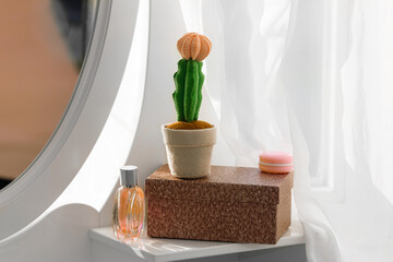 Felt Mondkaktus Gymnocalycium with a rose bud stands on a white dressing table on a brown  podium with a perfume bottle and a macaroon lip gloss.