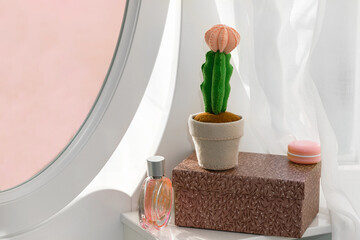 Felt Mondkaktus Gymnocalycium with a rose bud stands on a white dressing table on a brown  podium with a perfume bottle and a macaroon lip gloss.