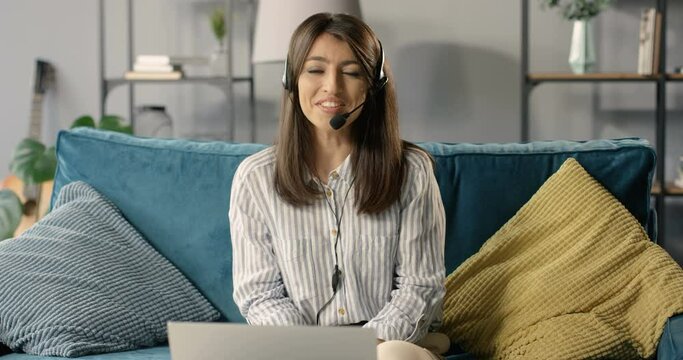Beautiful young happy woman blogger in headset and mic sitting on couch and talking to camera. Lesson online. Video blog recording. Female teacher or cauch teaching in internet. Vlogger speaking.