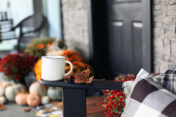 Steaming coffee cup sitting on arm of rocking chair on a front porch that has been decorated for autumn with heirloom white, orange and grey pumpkins and mums. Selective focus with blurred background.
