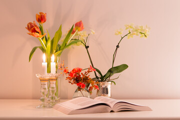 Book left open on table, with lighted candles, orchid, begonia and tulips in soft focus background seen in soft lamp light 