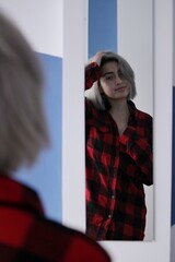 young girl with blonde hair standing in front of a mirror looking happy at herself wearing a red checkered shirt and touching her head
