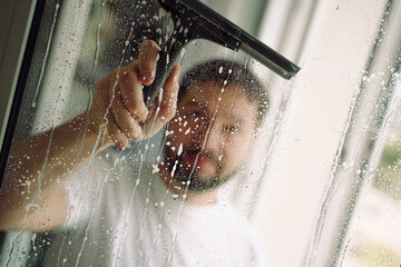 Young handsome bearded man doing spring cleaning in an apartment washing window with small...