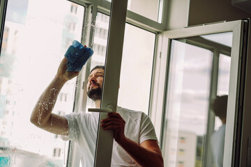 Young handsome bearded man doing spring cleaning in an apartment washing window with cloth