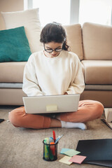 Charming lady in casual clothes sitting on the floor and using a laptop while doing homework and smile