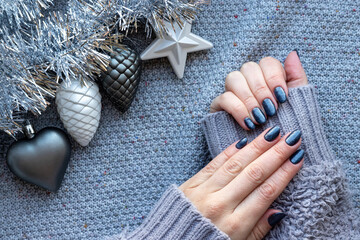 Female hands in gray knitted sweater with beautiful manicure - dark gray blue glittered nails on knitted background with silver tinsel garland and Christmas tree toys: heart, star and pine cones