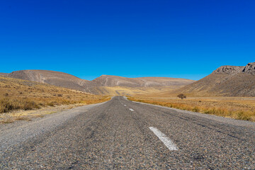 The road leads to the mountains. Mountain highway. Asphalt texture. Country highway. Mountains and hills in autumn. Mountain plants in autumn. Plants on the roadside. Dry yellow grass. Blue sky
