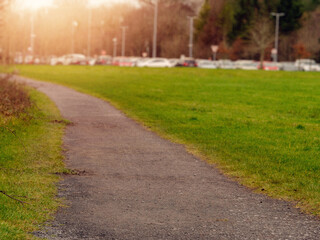 Path in a park in focus leads to busy car park full of cars out of focus in the background. Warm sunny day, Sun flare, Concept outdoors activity