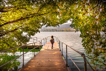 Avigliana, Italy. October 10th, 2020. Woman walks on the jetty of the lake, framed through the branches showing autumn foliage.