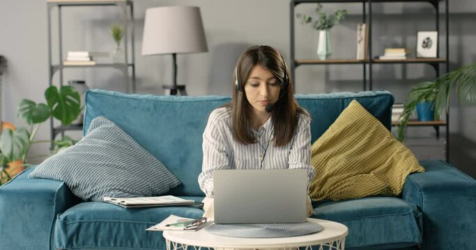 Beautiful woman in headset sitting on couch, videochating on laptop computer and working. Female teacher or cauch teaching online. Freelancer working at home via webcam with videochat and talking.