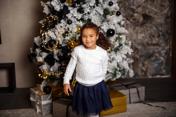 Happy smiling mixed girl with dark hair sitting on the carpet. Christmas tree in the background.