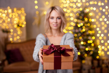 Christmas and New Year concept - beautiful blonde woman giving Christmas gift in decorated living room with Christmas tree and lights. Focus on gift box