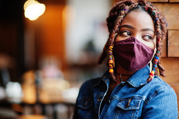 Stylish african american woman with dreadlocks afro hair, wear jeans jacket and face protect  mask at restaurant. New normal life after coronavirus epidemic.