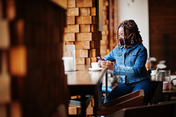 Stylish african american woman with dreadlocks afro hair, wear jeans jacket and face protect  mask at restaurant, hold cellphone. New normal life after coronavirus epidemic.