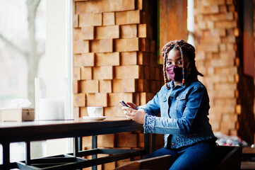 Stylish african american woman with dreadlocks afro hair, wear jeans jacket and face protect  mask at restaurant, hold cellphone. New normal life after coronavirus epidemic.