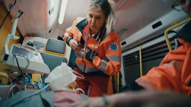 Female and Male EMS Paramedics Provide Medical Help to an Injured Patient on the Way to a Healthcare Hospital. Emergency Care Assistant Inspect the Victim with a Flashlight in an Ambulance.