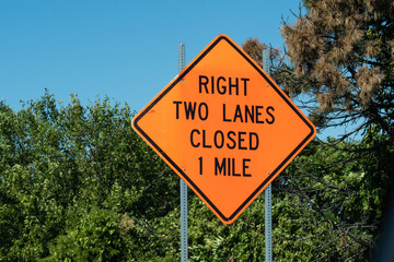 Bright orange Diamond shape sign ways "right two lanes closed 1 mile" on metal post with