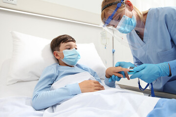 Nurse with pulse oximeter on patient child in hospital bed, wearing protective visor mask, corona virus covid 19 protection concept 