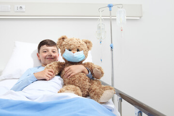 Smiling child in hospital bed with teddy bear wearing protective mask, corona virus covid 19 protection concept 