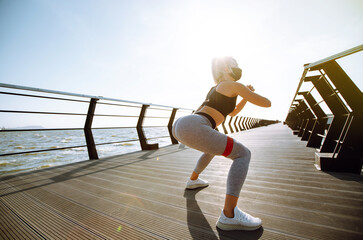 Young woman in protective  mask and sports outfit doing exercises outdoors in the morning. Sport woman  doing stretching exercise near the sea. Sport, Active life, Jogging during quarantine. Covid-19.