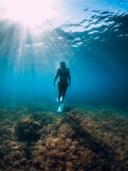 Freediver woman with white fins glides over seaweed bottom with sun rays. Freediving underwater in sea