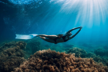 Freediver woman with white fins glides over seaweed bottom with sun rays. Freediving underwater in sea
