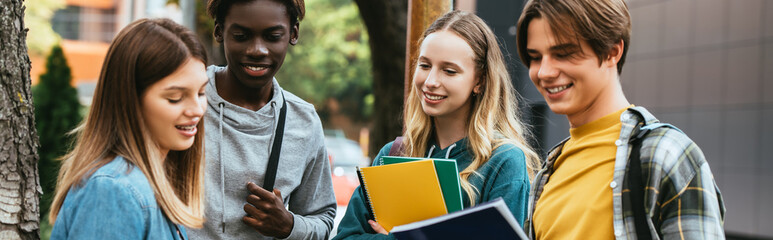 Fototapeta premium Horizontal crop of smiling multiethnic teenagers with notebooks sanding near tree outdoors