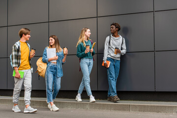 Smiling multiethnic teenagers with coffee to go and notebooks walking near building outdoors