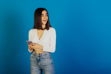 Happy smiling girl hold modern smart phone in hands, is smiling and looking away standing isolated on blue background with copy space.