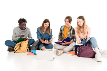 Smiling multicultural teenagers writing on notebooks near laptop on white background