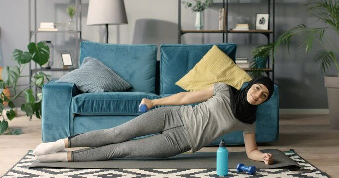 Young beautiful Arabian woman in hijab standing in side plank on yoga mat and doing exercise with dumbbell for arm biceps. At home. Pretty slim sporty muslim female balancing and working out on floor.
