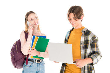 Smiling teenager holding laptop near friend with notebooks talking on smartphone isolated on white