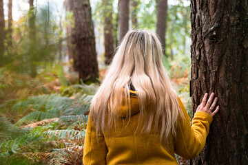 Young blonde woman in yellow coat leaning on a tree in the autumn forest	
