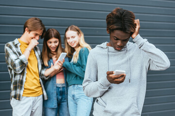 Selective focus of upset african american boy using smartphone near smiling teenagers with cellphone outdoors