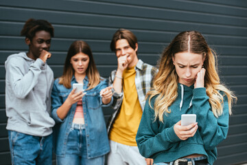 Selective focus of upset girl using smartphone near laughing multiethnic teenagers outdoors