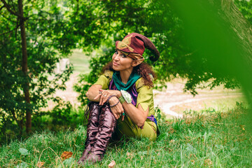 Sly look of a wood elf sitting on the grass in a magic forest. A woman dressed as an elv, she has elven ears and an elven hat.Carnival and costume party.
