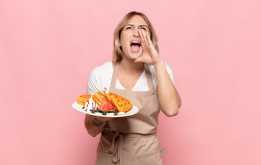 young blonde woman feeling happy, excited and positive, giving a big shout out with hands next to mouth, calling out