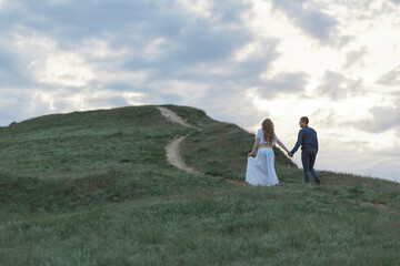 A couple in love, a pregnant wife in a white outfit are walking in a hilly field. Overcast weather