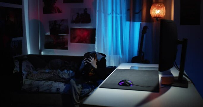 An upset and depressed desperate teenage boy sits on the bed and holds his head. Long shot. frontal